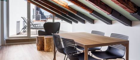 Un comedor moderno con una mesa de madera y sillas negras, un taburete de madera, y vistas a un paisaje urbano a través de amplios ventanales.