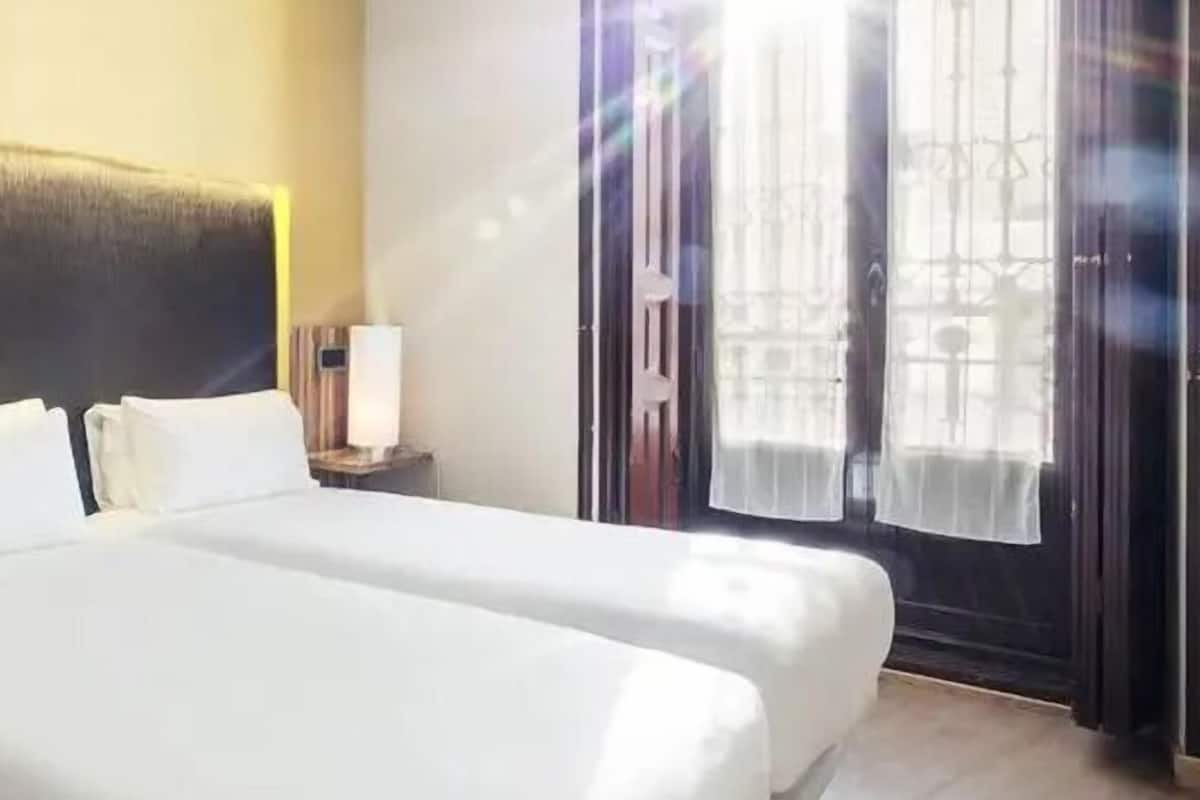 A hotel room with a large bed, a wooden headboard, a bedside table with a lamp, and a view of a building through the window.