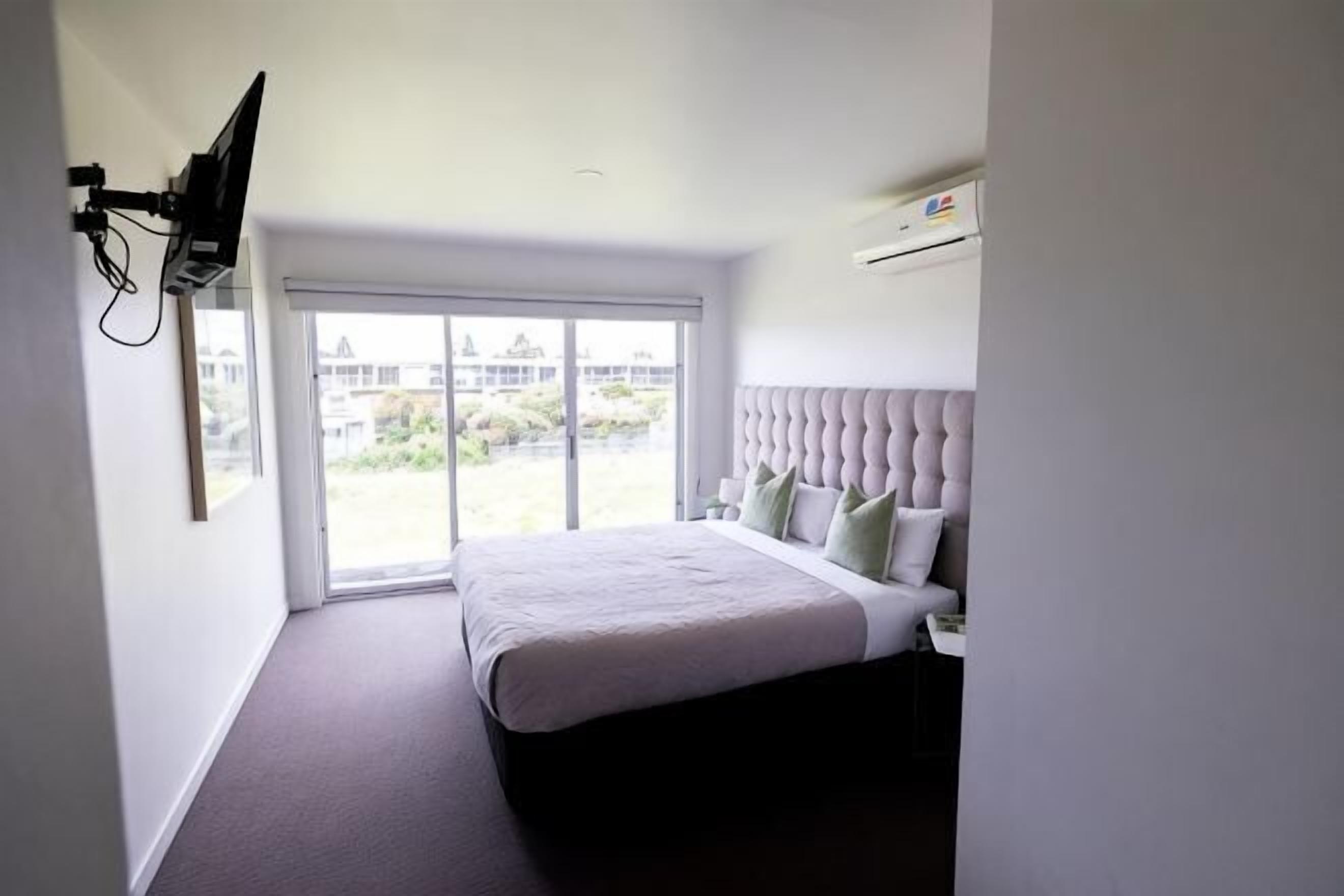 A bedroom with a bed, a television mounted on the wall, and a window with a view of buildings.