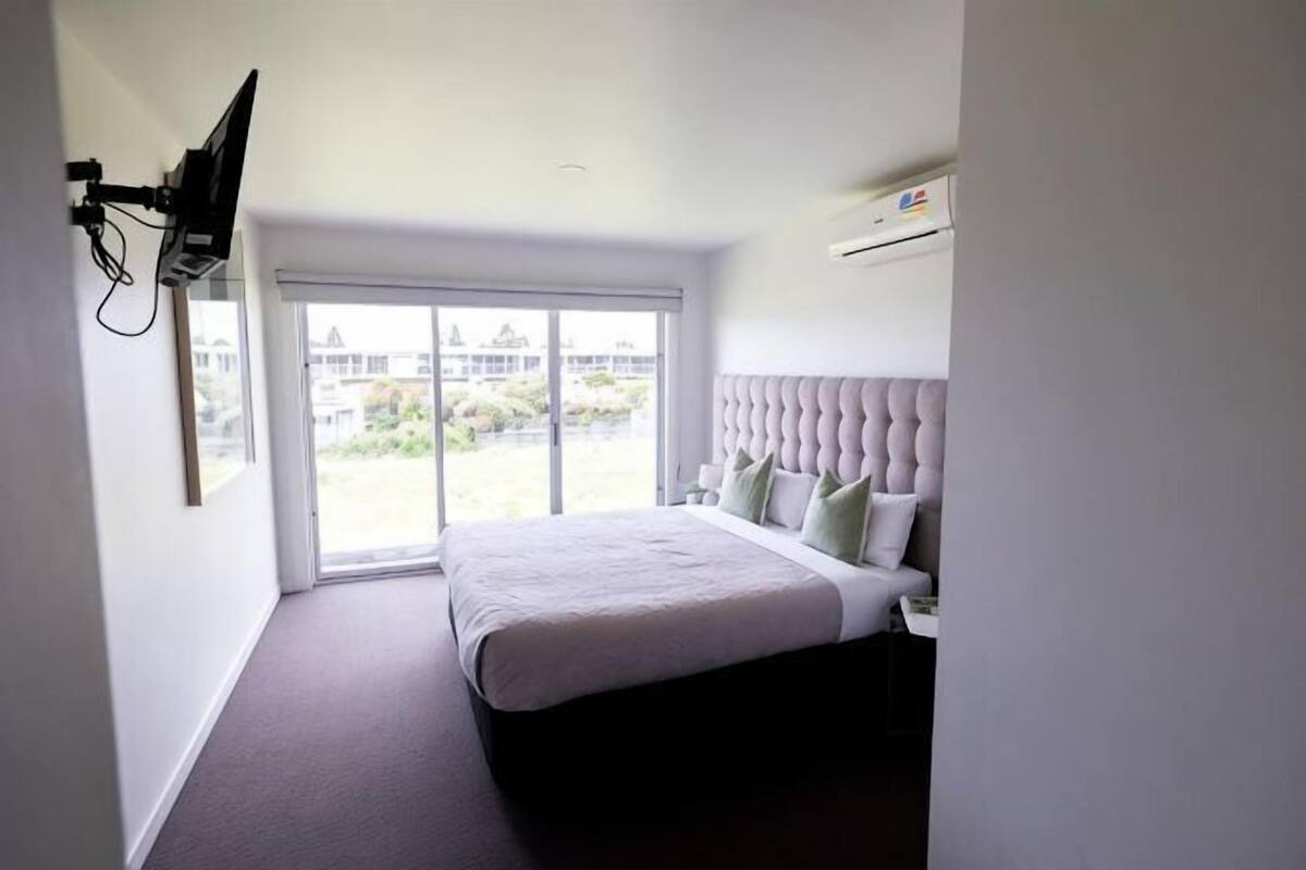 Un dormitorio con cama, televisor montado en la pared y ventana con vista a edificios.