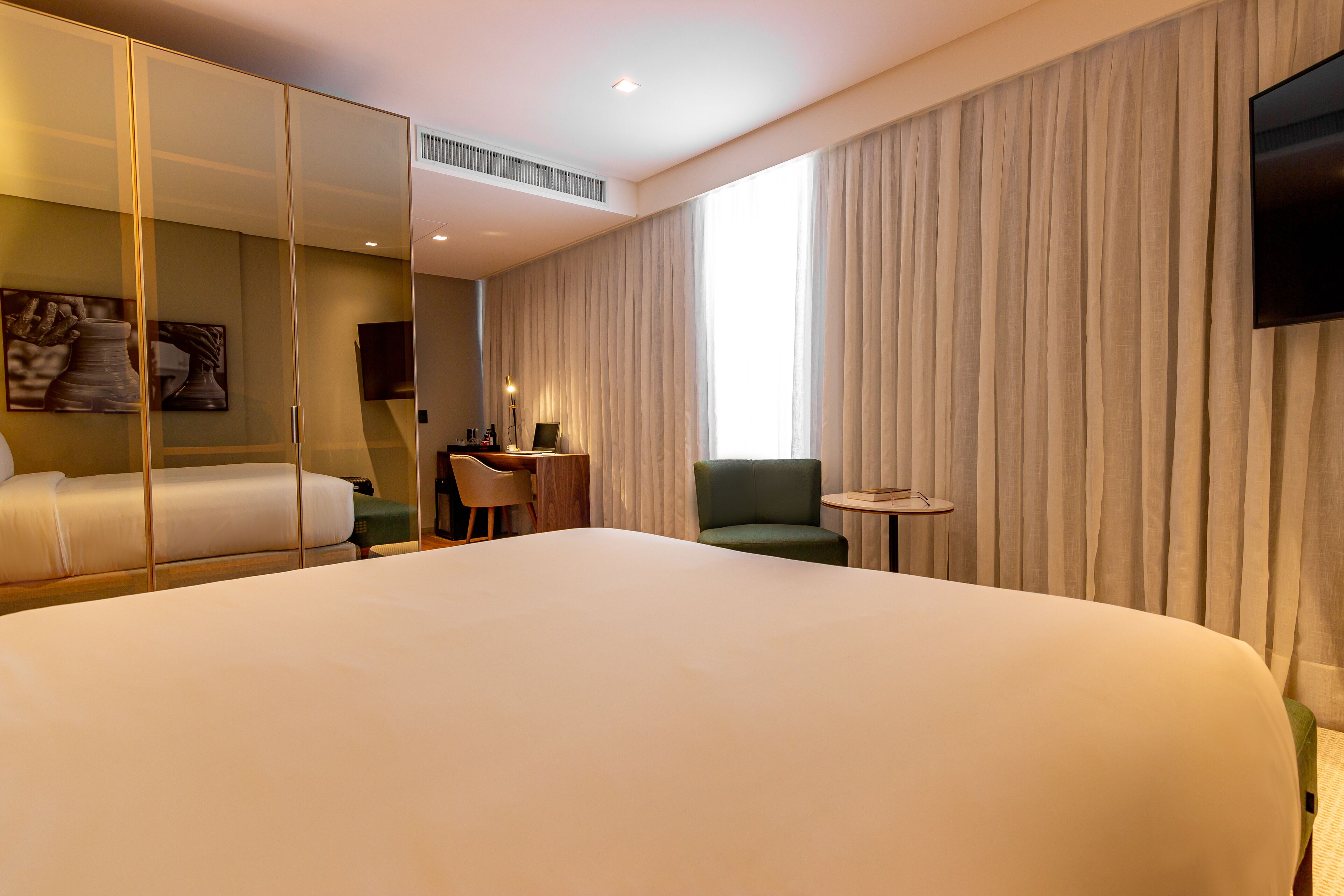 A hotel room with a large bed, a desk, a chair, a small table, a TV, and a large window with curtains.