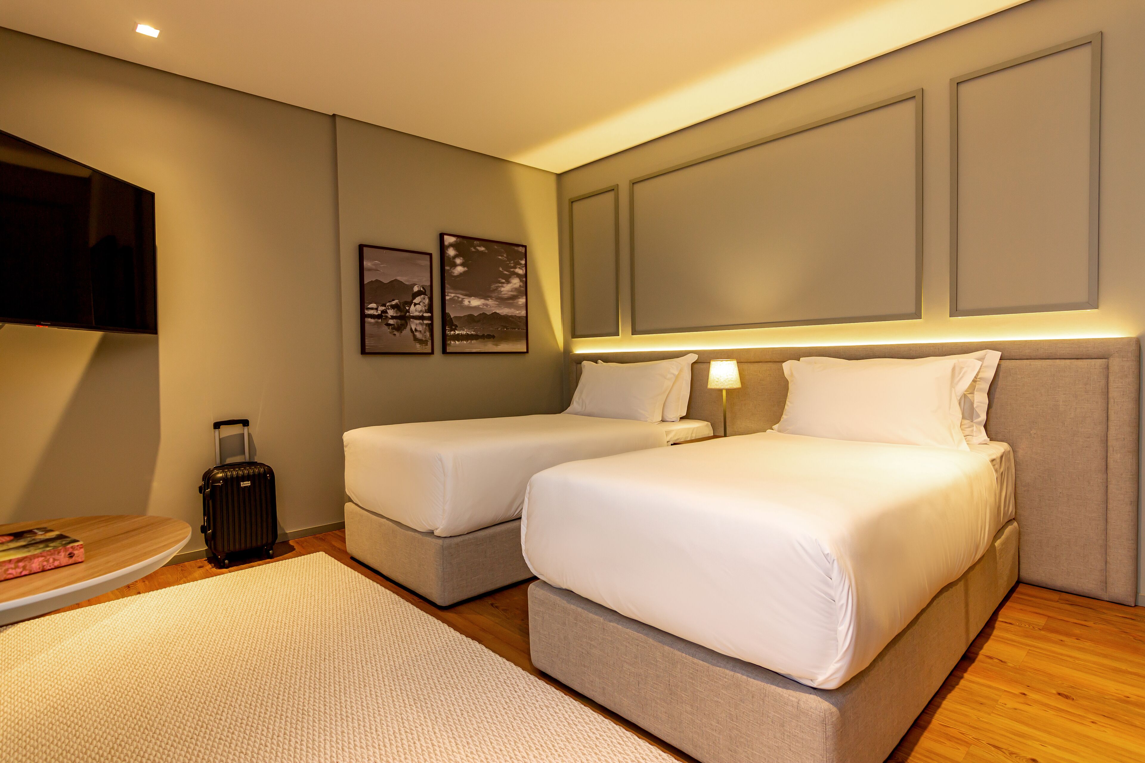 A hotel room with two beds, a TV, a suitcase, and a small table with a book.