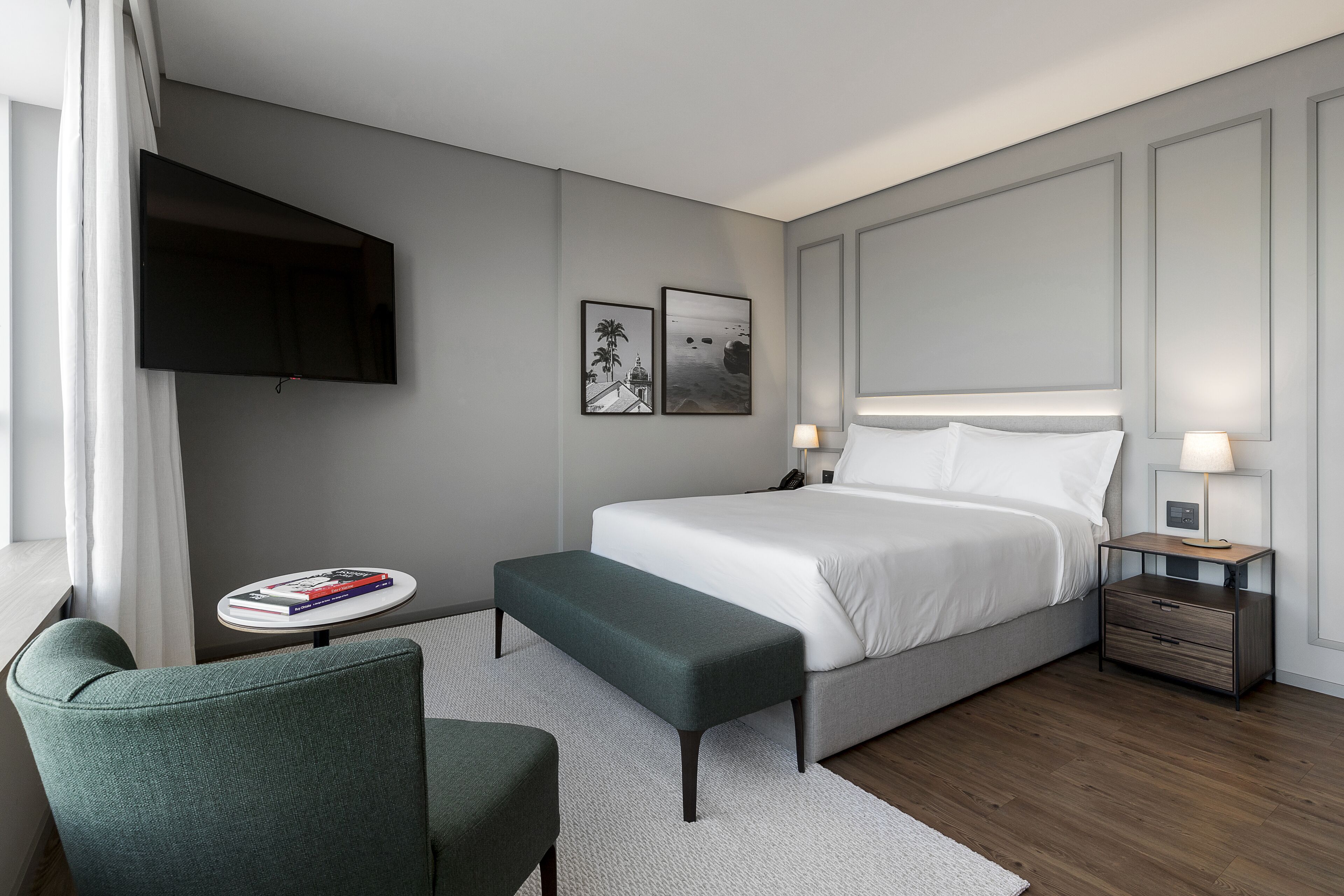 A modern hotel room with a large bed, bedside tables, a flat-screen TV mounted on the wall, and a small seating area with a chair and a bench.