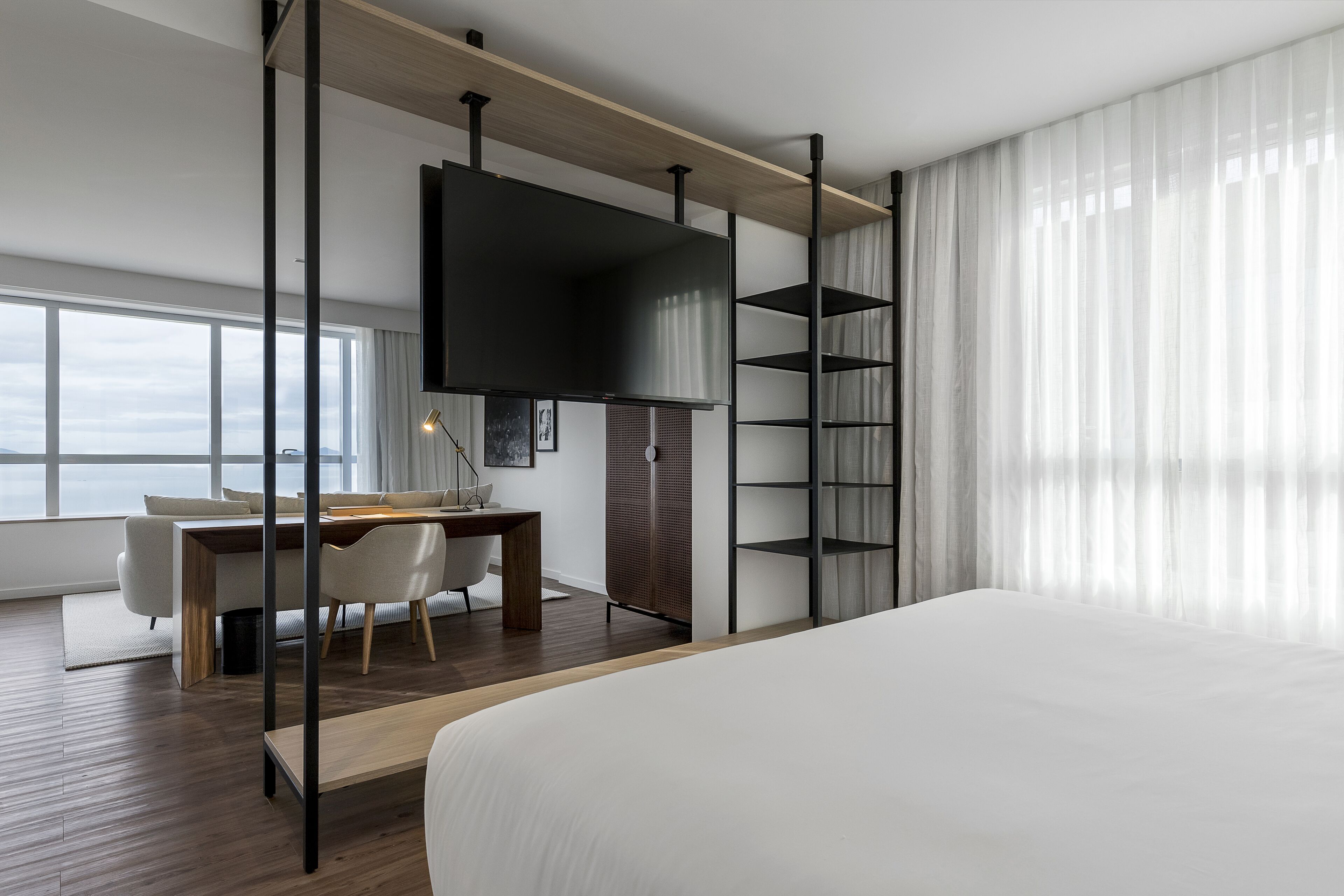 Quarto de hotel moderno com uma cama grande, uma escrivaninha com cadeira, uma TV fixada em uma prateleira de madeira e uma janela ampla com cortinas.