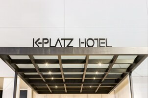 Exterior do Hotel K-Platz com design moderno, apresentando fachada de metal e vidro e uma placa chamativa sobre a entrada.