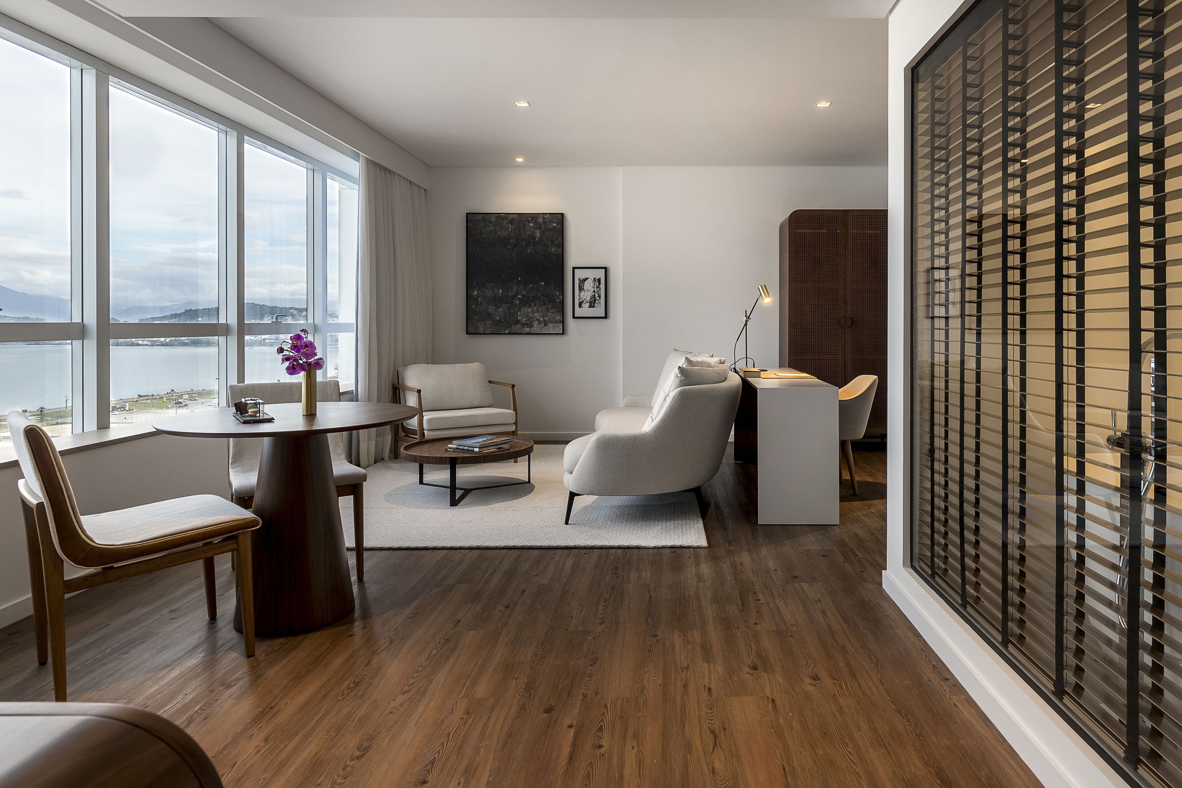 Una sala de estar moderna con vista al agua, una mesa redonda, un sofá y un amplio ventanal con persianas.