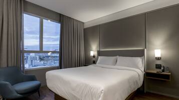 Una habitación de hotel con una cama grande, una silla, una mesita de noche con un teléfono y vistas a la ciudad a través de una ventana.