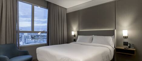 Una habitación de hotel con una cama grande, una silla, una mesita de noche con un teléfono y vistas a la ciudad a través de una ventana.