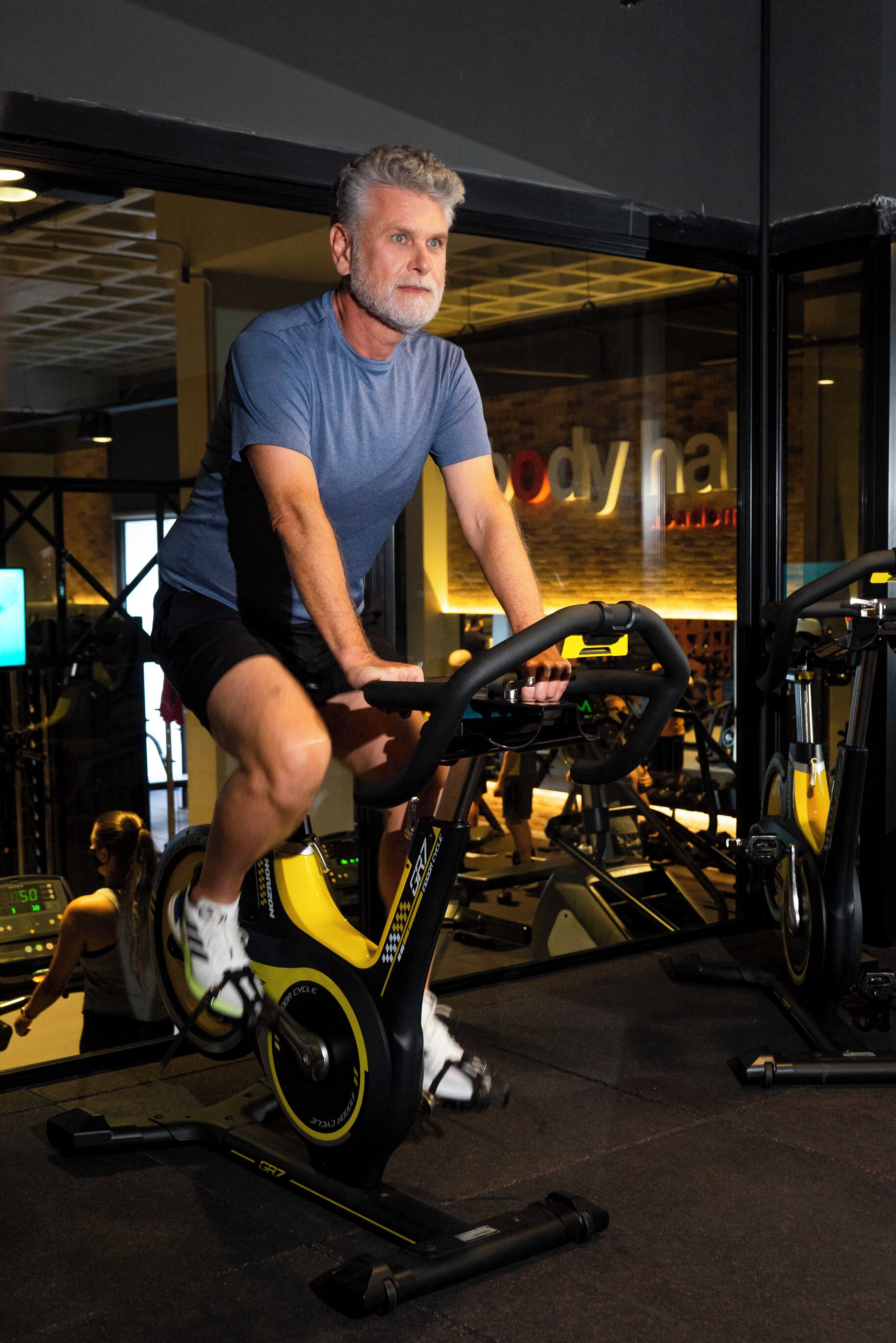 Ein Mann fährt auf einem Stillstandfahrrad in einem Fitnessstudio mit Trainingsgeräten und einem sichtbaren Logo an der Wand.