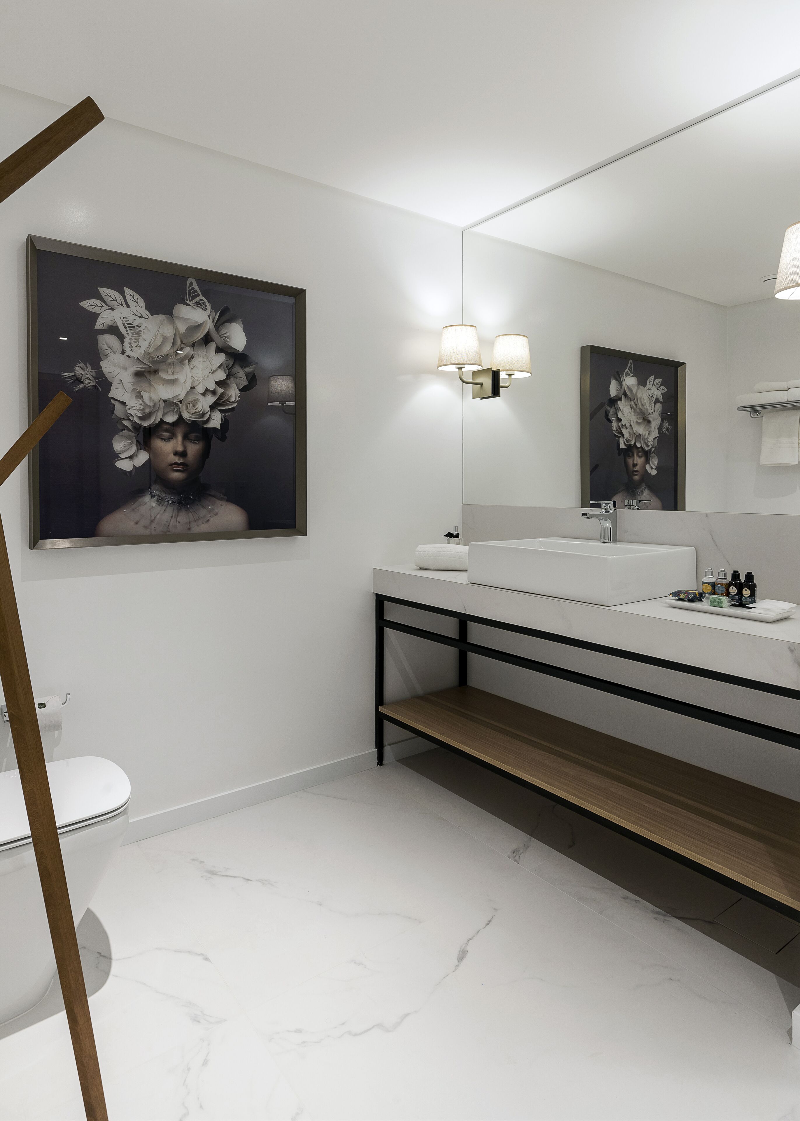 Un baño moderno con suelo de mármol blanco, un lavabo blanco, un espejo con marco negro y una gran obra de arte floral sobre el espejo.