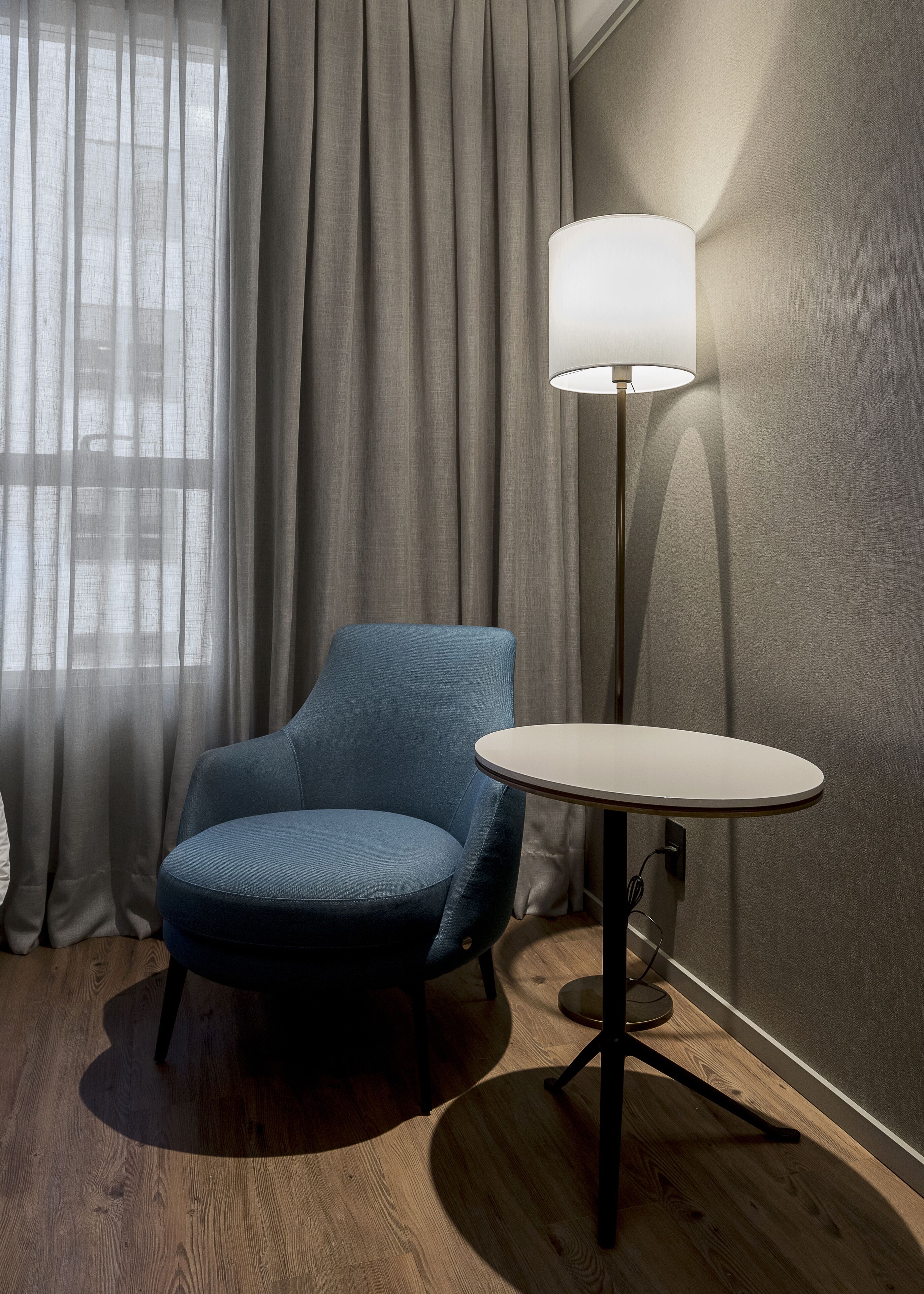 A room with a blue armchair, a round table, a floor lamp, and a window with curtains.