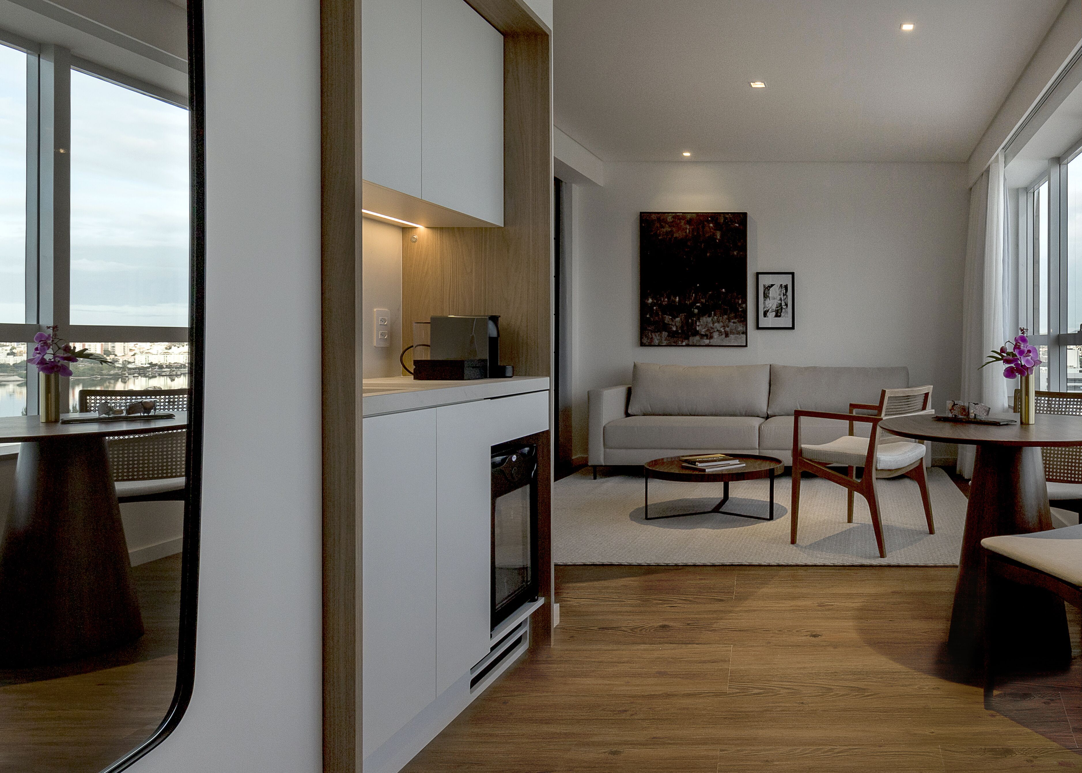 A modern living room with a sofa, coffee table, and dining table. There is a kitchen area with a stove and cabinets. The room has large windows with a view of the city.