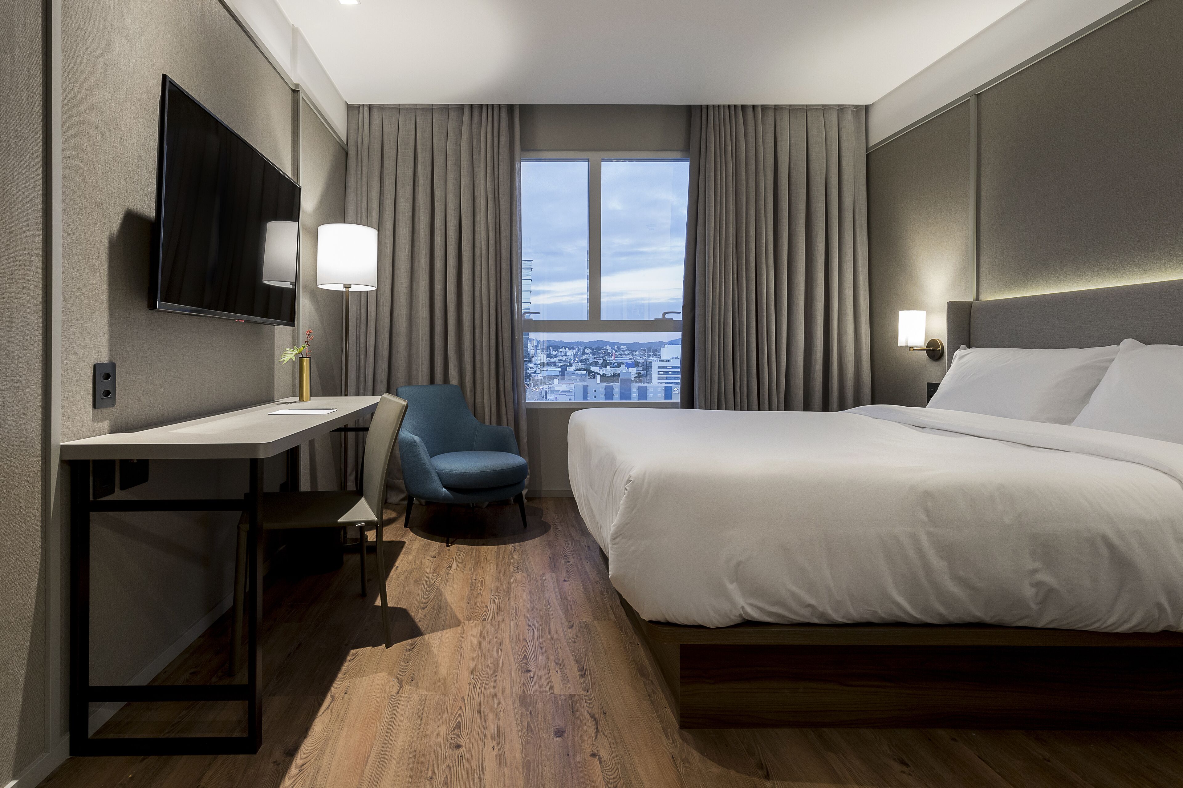 Une chambre d’hôtel équipée d’un lit, d’un bureau, d’une chaise, d’une télévision et d’une fenêtre donnant sur la ville.