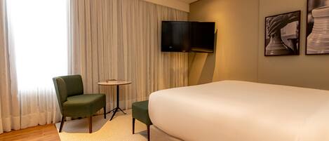 A hotel room with a large bed, a green armchair, a small table, and a flat-screen TV mounted on the wall.