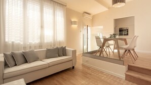 Ein modernes Wohnzimmer mit Sofa, Esstisch, Stühlen und einem durch eine Glastrennwand sichtbaren Küchenbereich.