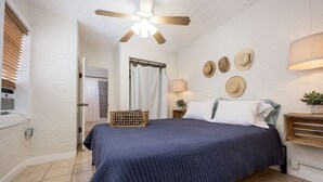 A bedroom with a bed, a ceiling fan, a window with blinds, a door, a wall-mounted lamp, and a wall with hats. - The Blue House (Truth or Consequences)