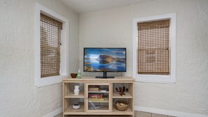 A corner room with a TV on a wooden stand, two windows with blinds, and a tiled floor. - The Blue House (Truth or Consequences)