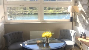 A room with a large window overlooking a river and forest, a round glass table with a vase of yellow flowers, two grey armchairs, and a lamp on a wooden table. - Country house for 15 people, waterfront in the Ardennes nature park (Revin)