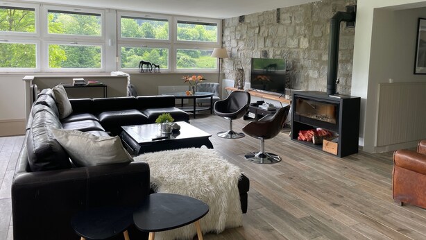 A modern living room with a stone wall, large windows, a fireplace, and a cozy seating area. - Country house for 15 people, waterfront in the Ardennes nature park (Revin)