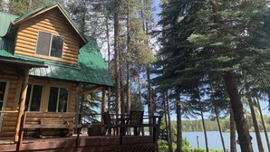 Une cabane en rondins avec un toit végétalisé, une véranda meublée en bois et une vue sur un lac entouré d’arbres.