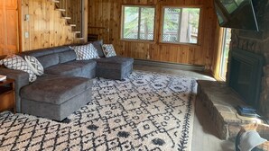 Un intérieur de chalet chaleureux avec un canapé d’angle, un tapis à motifs et une cheminée en pierre.