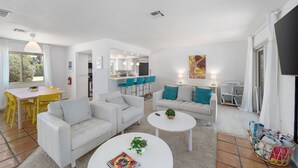 Ein modernes Wohnzimmer mit weißen Sofas, einem Esstisch mit gelben Stühlen, einer Küchenzeile und Blick auf Grünfläche.