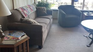 Un salón acogedor con sofá, sillón y una mesita baja con revistas. Amplias ventanas ofrecen vistas a un lago y barcos.