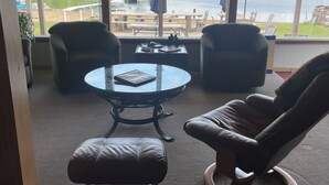 Habitación con un ventanal grande que da a un lago y montañas. Hay una mesa redonda con sillas y un reposapiés de cuero.