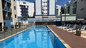A swimming pool with a wooden deck and lounge chairs, surrounded by modern buildings.