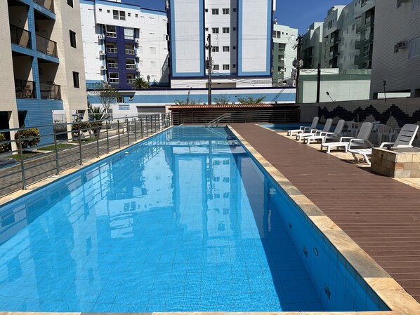 A swimming pool with a wooden deck and lounge chairs, surrounded by modern buildings.