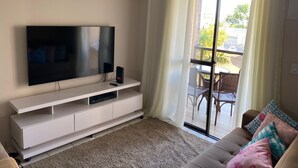 A living room with a flat-screen TV on a white TV stand, a brown sofa with patterned cushions, and a sliding glass door leading to an outdoor area with a table and chairs.
