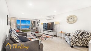 Una sala de estar moderna con un televisor grande, asientos cómodos, vista al mar y un reloj en la pared.