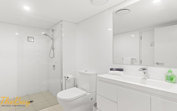 A modern bathroom with a glass-enclosed shower, a toilet, a sink with a mirror, and a green bottle of hand soap. - Shoal Bay Apartment one street back from the beach with water views (Shoal Bay)