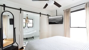 A bedroom with a bed, a ceiling fan, a mounted TV, a large window with curtains, and a door leading to a bathroom. - HISTORIC DOWNTOWN LOFT (Sevierville)