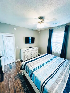 Une chambre à coucher comprenant un lit, un ventilateur de plafond, une télévision, une commode et une fenêtre avec des rideaux.