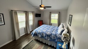 Ein Schlafzimmer mit einem Bett, einem Deckenventilator, einem Fernseher und einem Fenster mit Vorhängen.