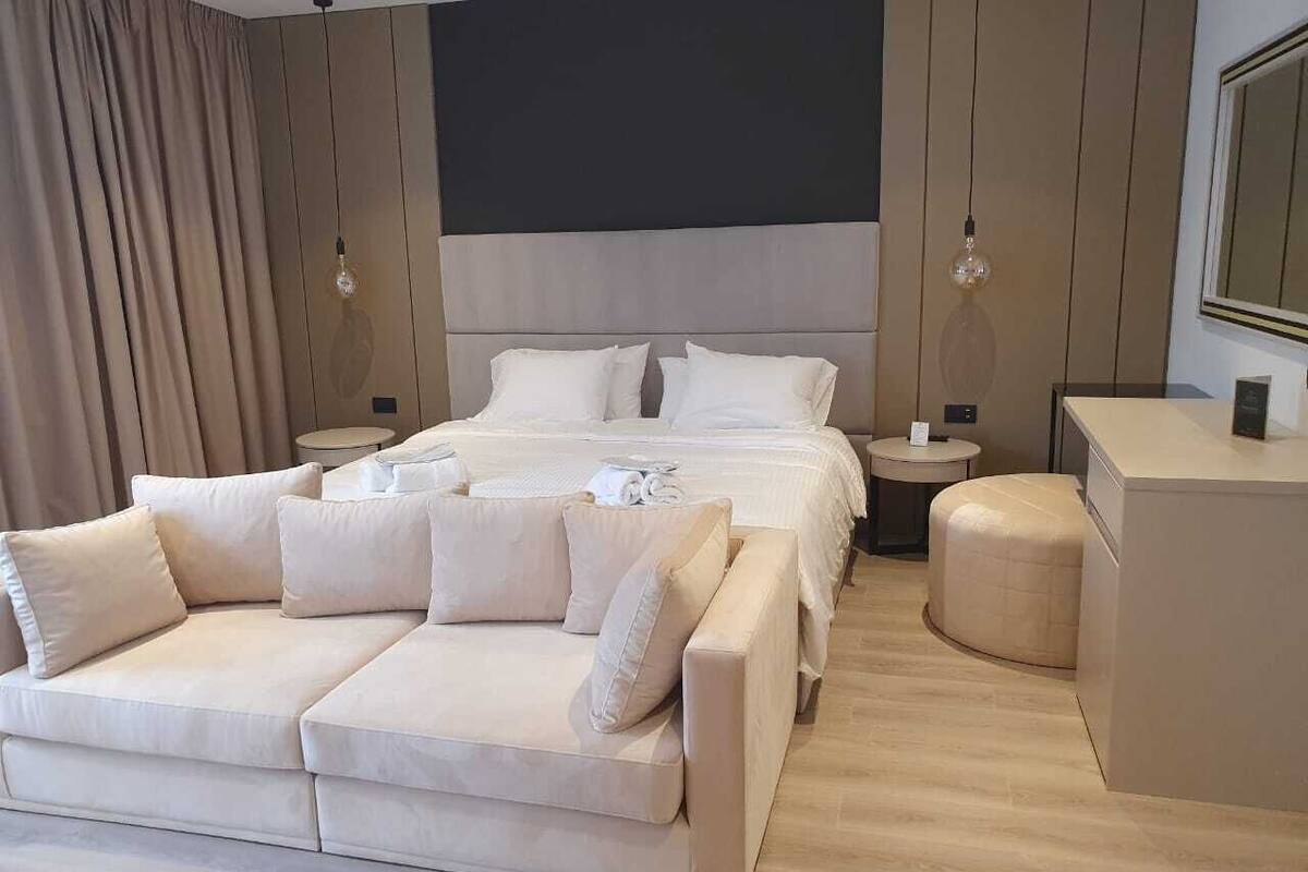 A modern bedroom with a large bed, a sofa, a round ottoman, and a small table with a lamp.