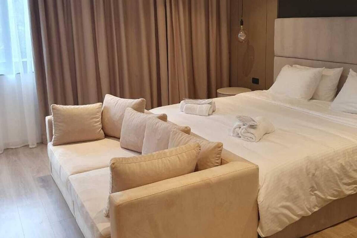 A modern hotel room with a large bed, two sofas, and a large window with curtains.