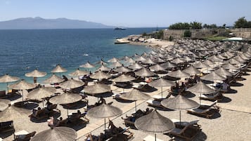 A beach with numerous sun loungers and thatched umbrellas, a clear view of the sea, and distant mountains.