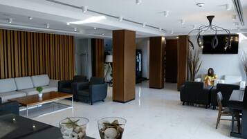 A modern hotel lobby with a sofa, armchairs, a coffee table, and a dining area with a table and chairs.