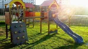 Un parc infantil amb elements d'escalada i tobogan, ubicat en un camp de gespa amb edificis al fons.