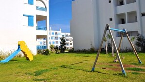 A playground with swings and a slide in front of a white building with blue accents.