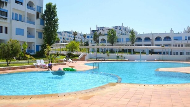 A swimming pool with a tiled deck, surrounded by buildings with balconies and a clear sky.