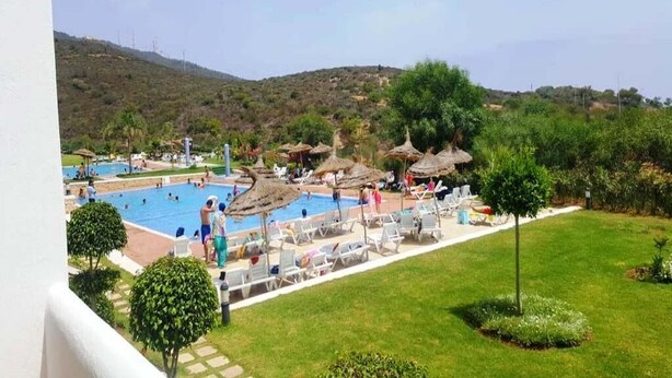 Ein Poolbereich mit Liegestühlen und Sonnenschirmen, umgeben von viel Grün und Hügeln im Hintergrund.