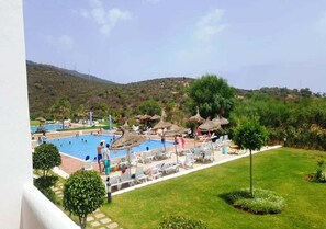 A pool area with lounge chairs and umbrellas, surrounded by greenery and hills in the background.