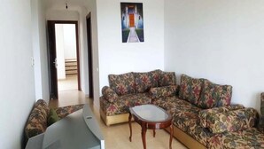 Ein Wohnzimmer mit einer Couch, einem Couchtisch und einem Fernseher. An der Wand hängt ein Gemälde, und eine Tür führt in einen anderen Raum.