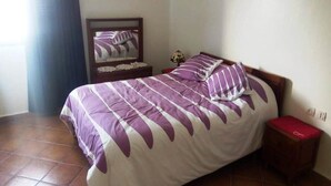 Een slaapkamer met een bed, nachtkastje, spiegel en een rode kruk.