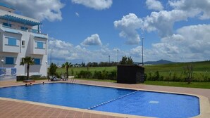 A swimming pool with a tiled deck, a building with blue accents, and a grassy landscape with mountains in the background. - Residence Smir Park Beach 1672 (Mdiq)