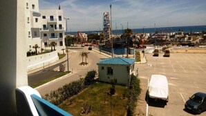 A parking lot with a covered car, a small building with a blue roof, and a large white building with balconies. - Residence Smir Park Beach 1673 (Mdiq)
