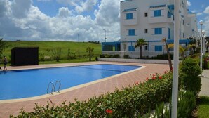 Une piscine dotée d’une terrasse carrelée, entourée d’un jardin bien entretenu et bordée d’un bâtiment à la façade blanche et aux accents bleus.